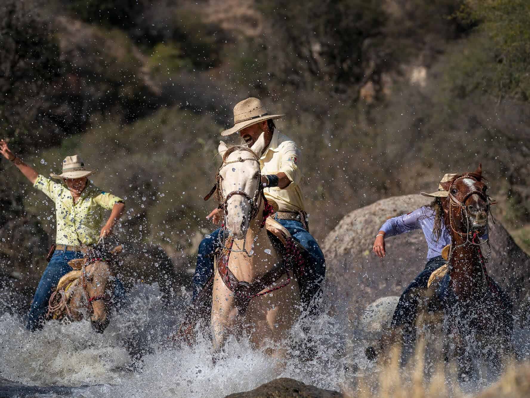 Jinetes con sombrero cruzando a caballo un arroyo entre rocas.
