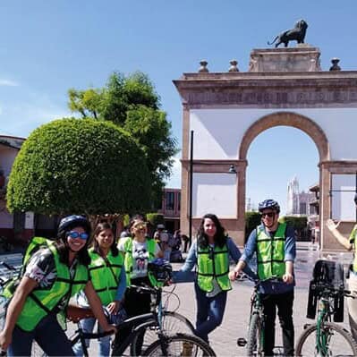 Grupo de turistas en bicicleta en Guanajuato, México, con monumento histórico de fondo.