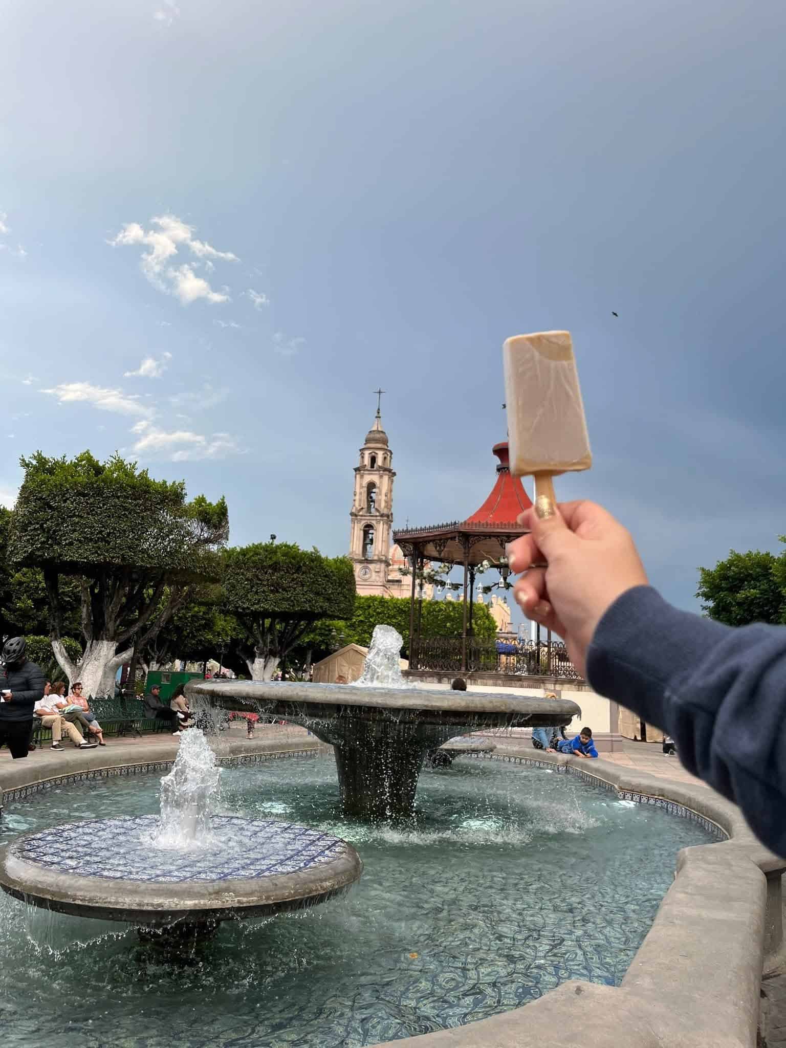 Persona disfrutando un helado en una plaza con fuente y iglesia en el fondo.