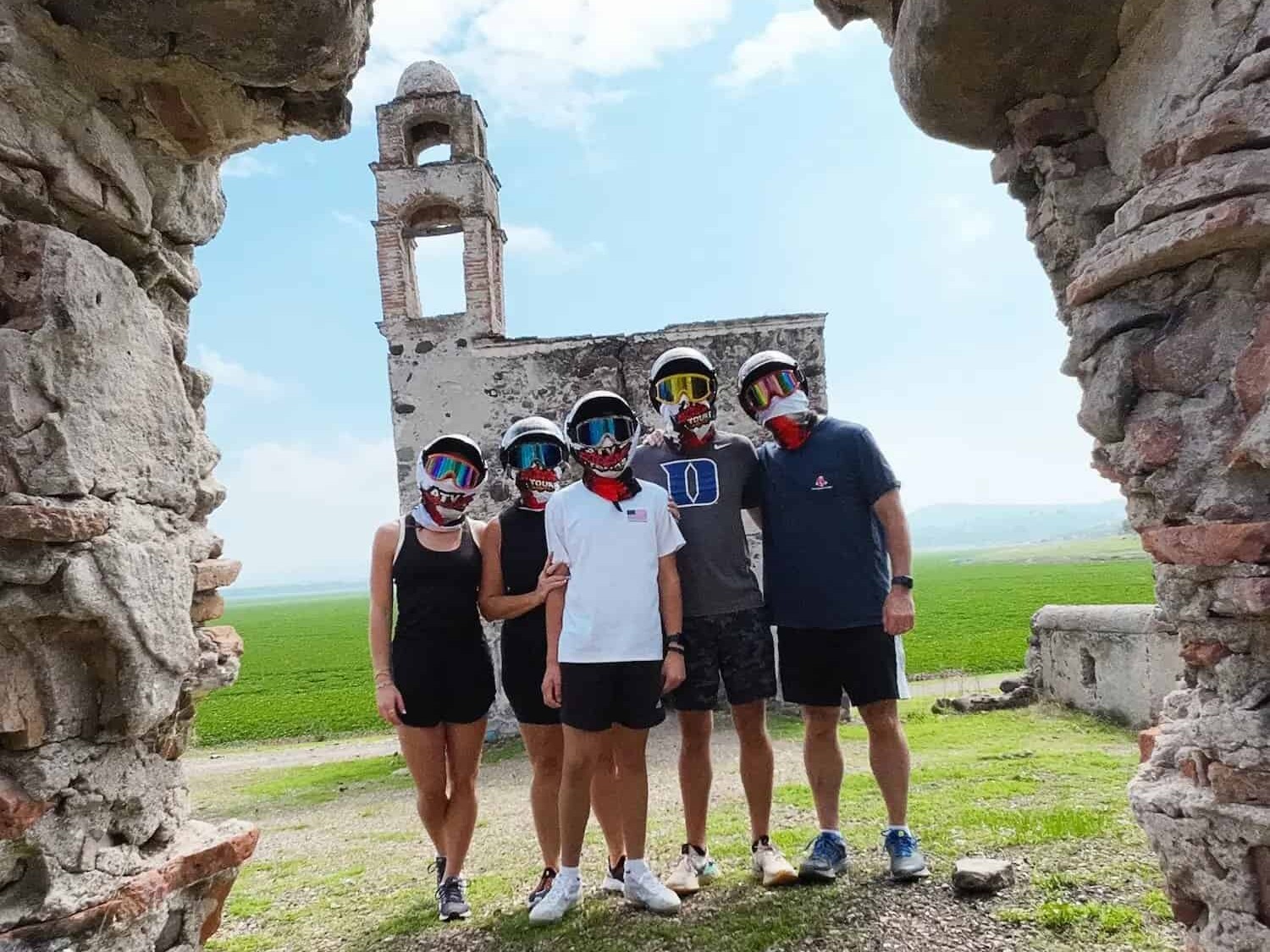 Group of helmeted travelers under a stone arch, with the ruins of a chapel in the background, on an ATV tour.