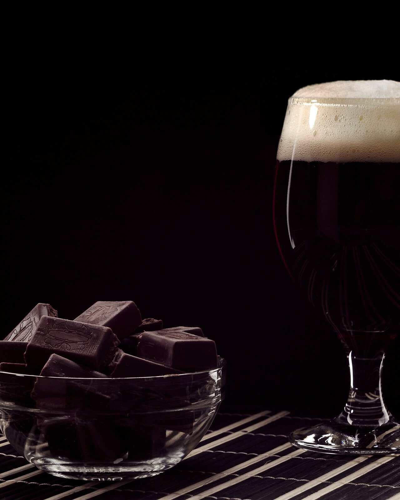 Copa de cerveza oscura junto a un tazón con trozos de chocolate amargo.