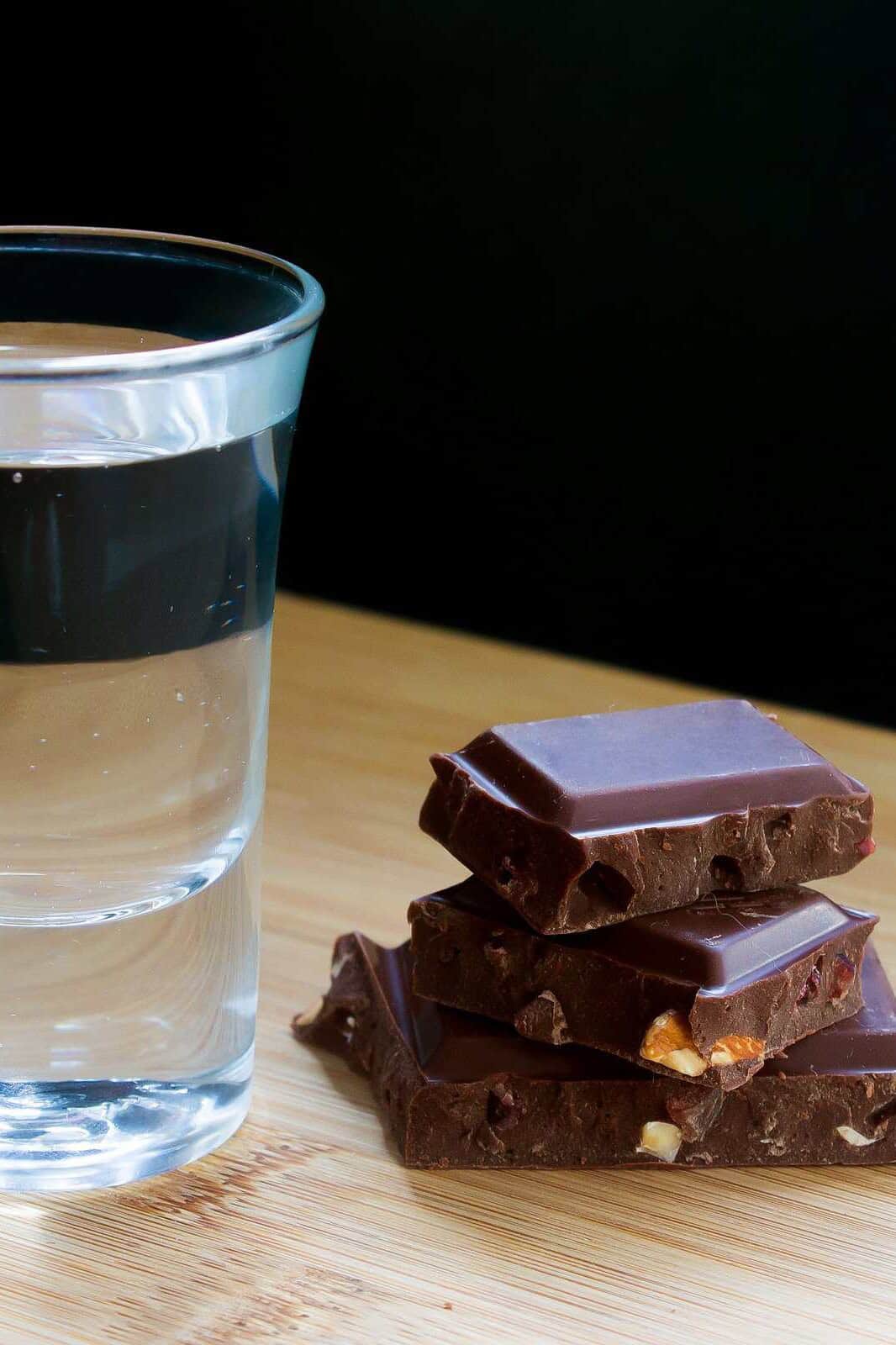 Vaso tequilero con destilado junto a trozos de chocolate amargo sobre una mesa de madera.
