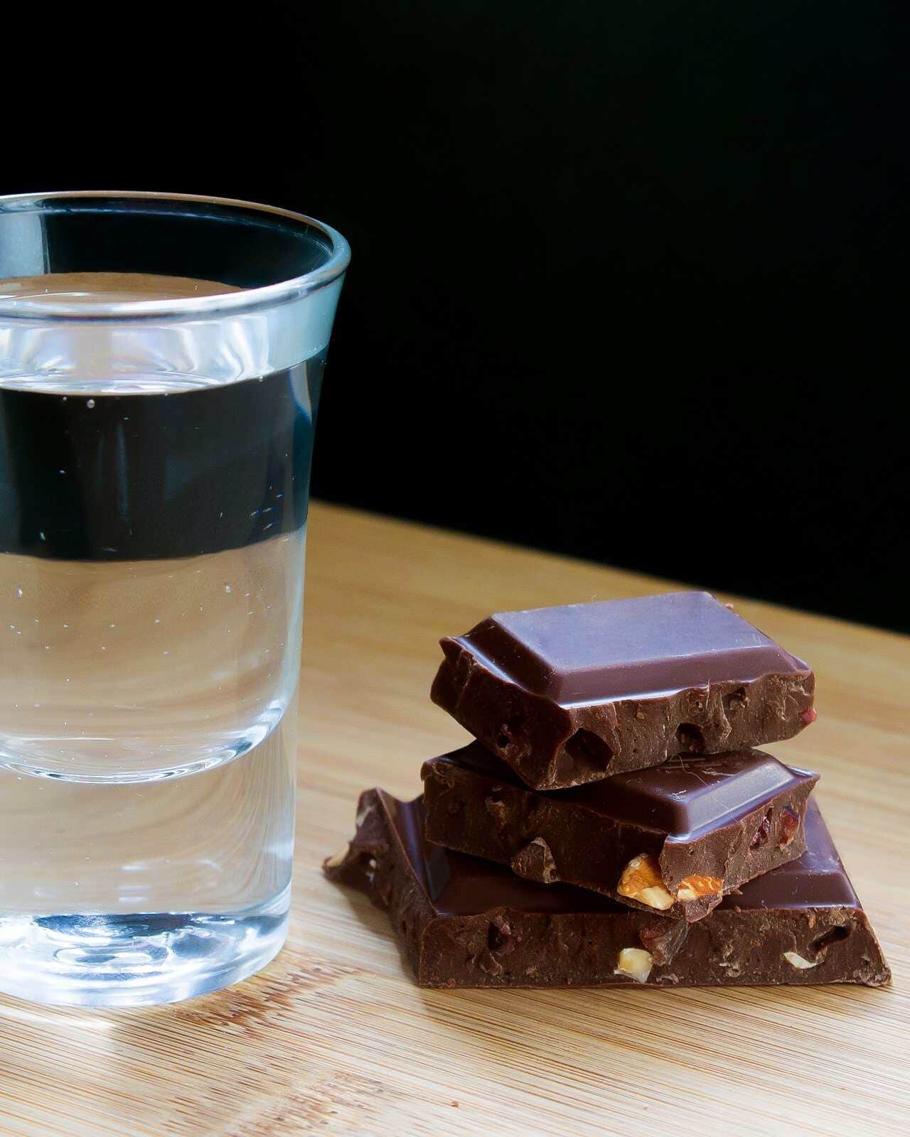 Vaso tequilero con destilado junto a trozos de chocolate amargo sobre una mesa de madera.