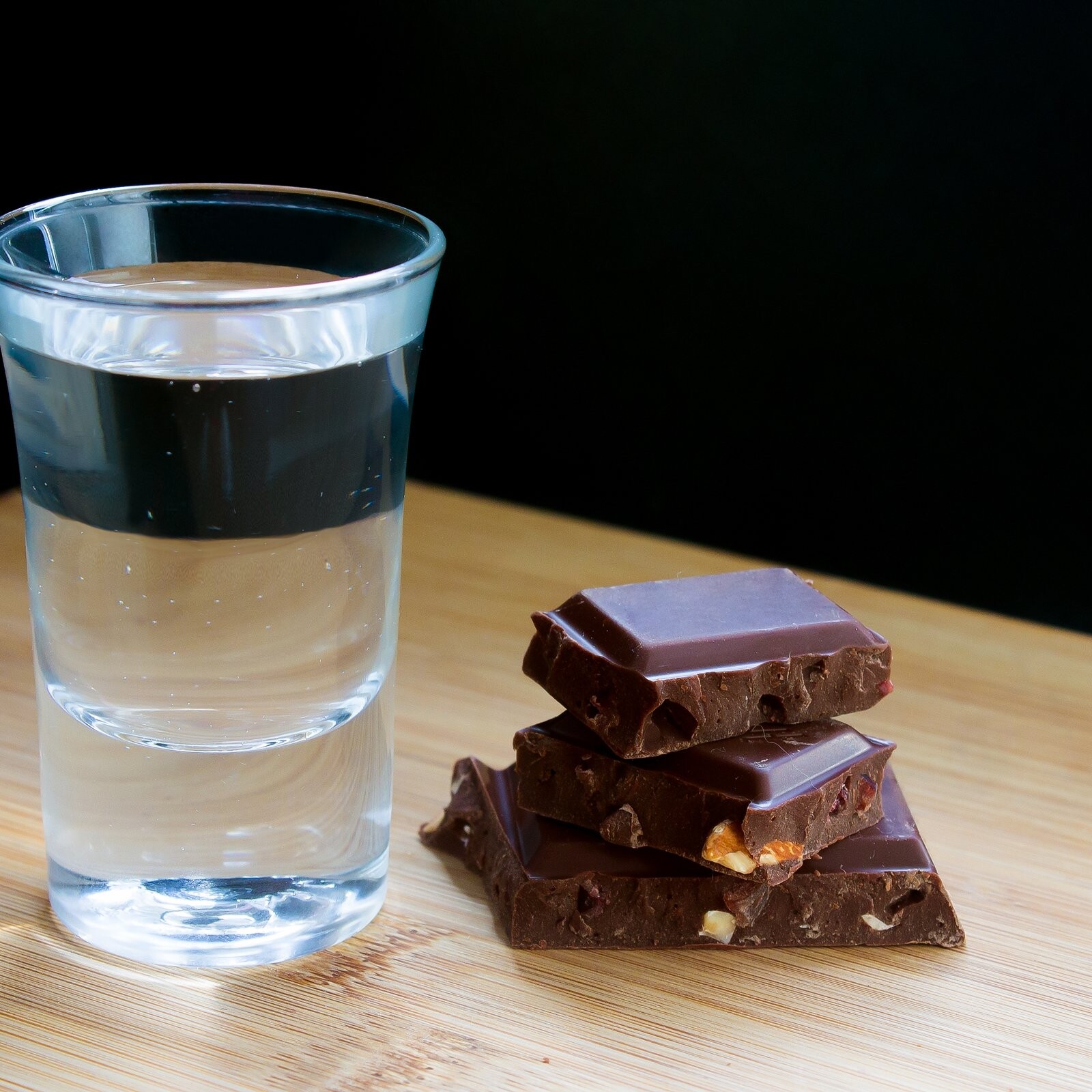 Vaso tequilero con destilado junto a trozos de chocolate amargo sobre una mesa de madera.