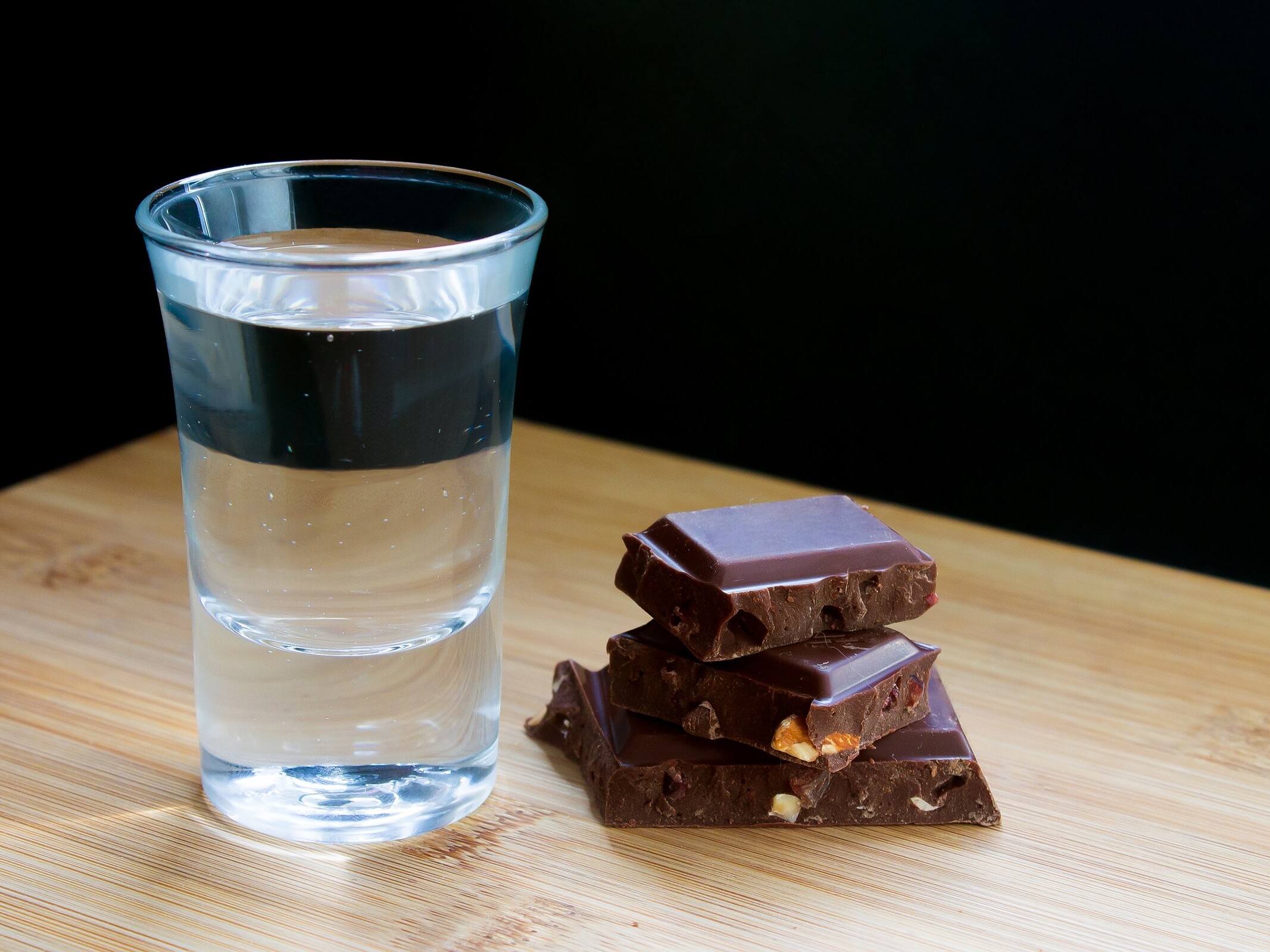 Vaso tequilero con destilado junto a trozos de chocolate amargo sobre una mesa de madera.