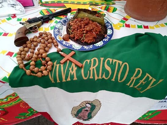 Table with a cristera flag «Viva Cristo Rey» with the Virgin of Guadalupe, a rosary, a wooden cross, an old pistol and a plate of typical food.