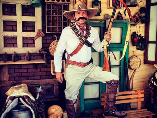Guide dressed as a Cristero fighter, with a sombrero, bandoliers, and a rifle, in a room with antique objects.