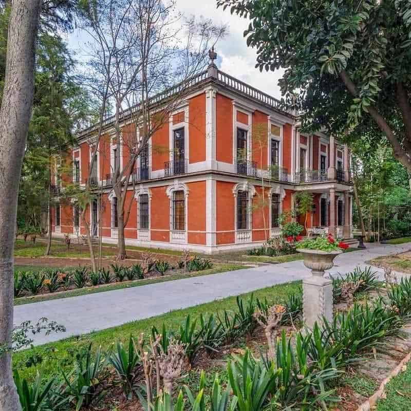 Red-fronted mansion surrounded by gardens and trees.