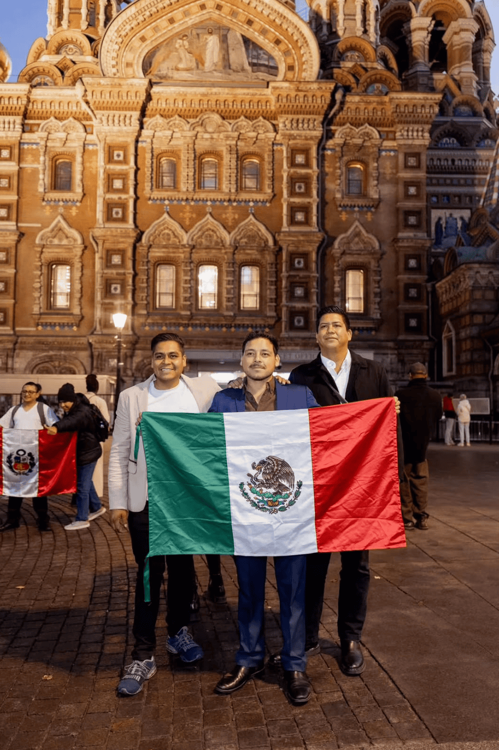 Tres jóvenes mexicanos sostienen la bandera de México frente a una iglesia durante un evento.