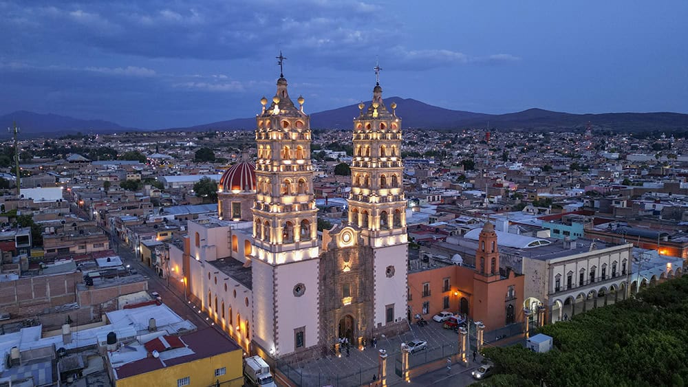 Catedral de Guanajuato iluminada al atardecer con vista urbana y monta&ntilde;as de fondo.