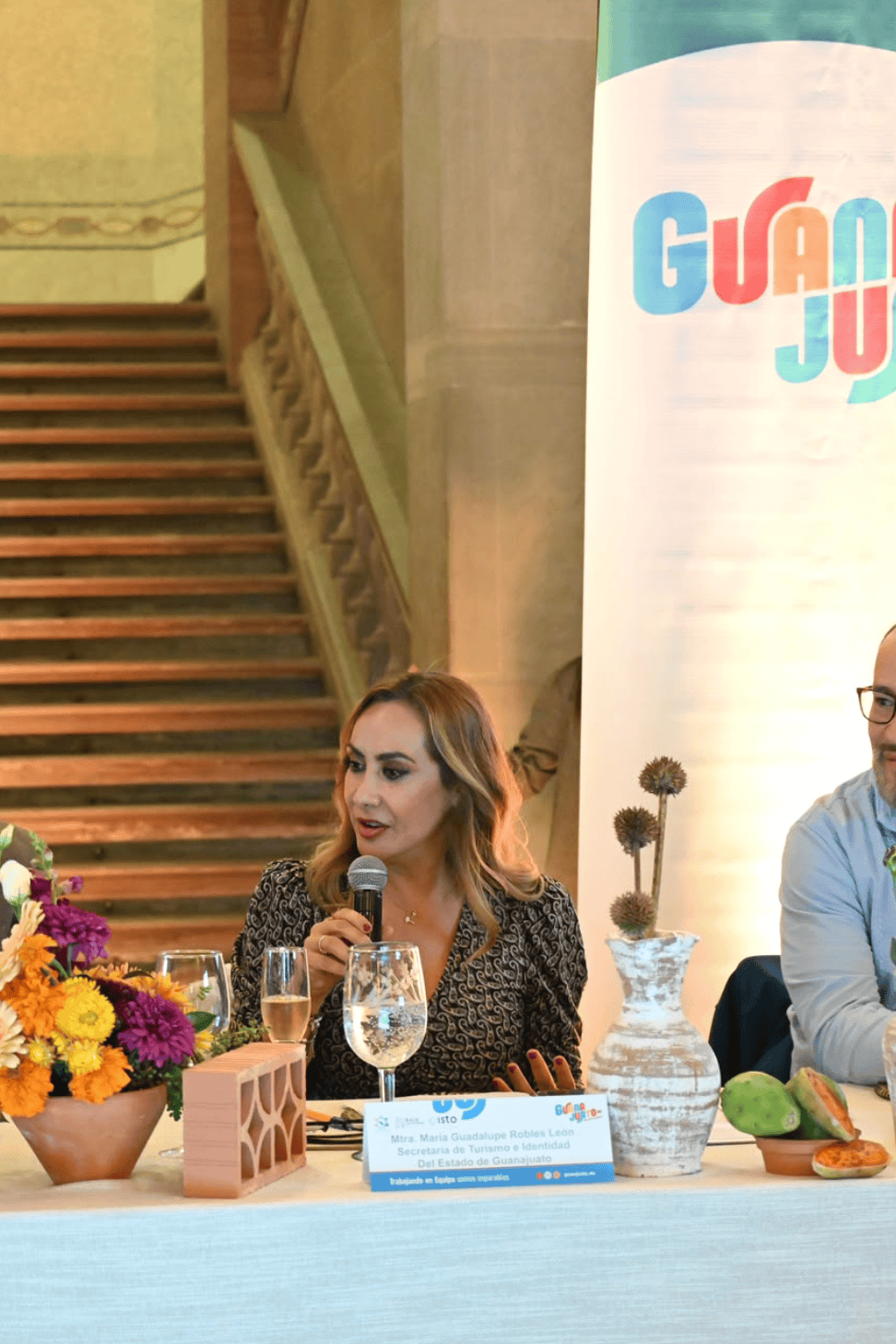 Mujer hablando en evento de turismo en Guanajuato con flores y cartel de fondo.
